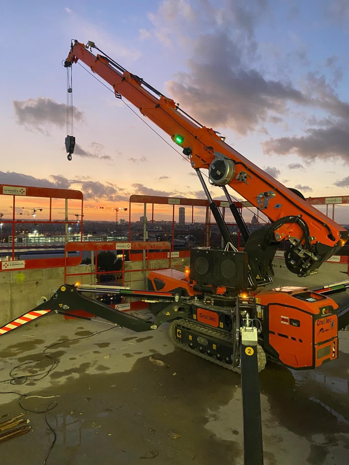 Mini-grue orange sur chenilles sur un toit de chantier lors d'un coucher du soleil.